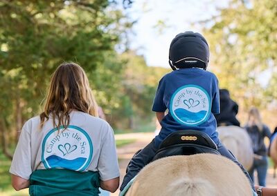 Child riding a horse with volunteer buddy at Camp By The Sea. Photo by Melissa Knowles.